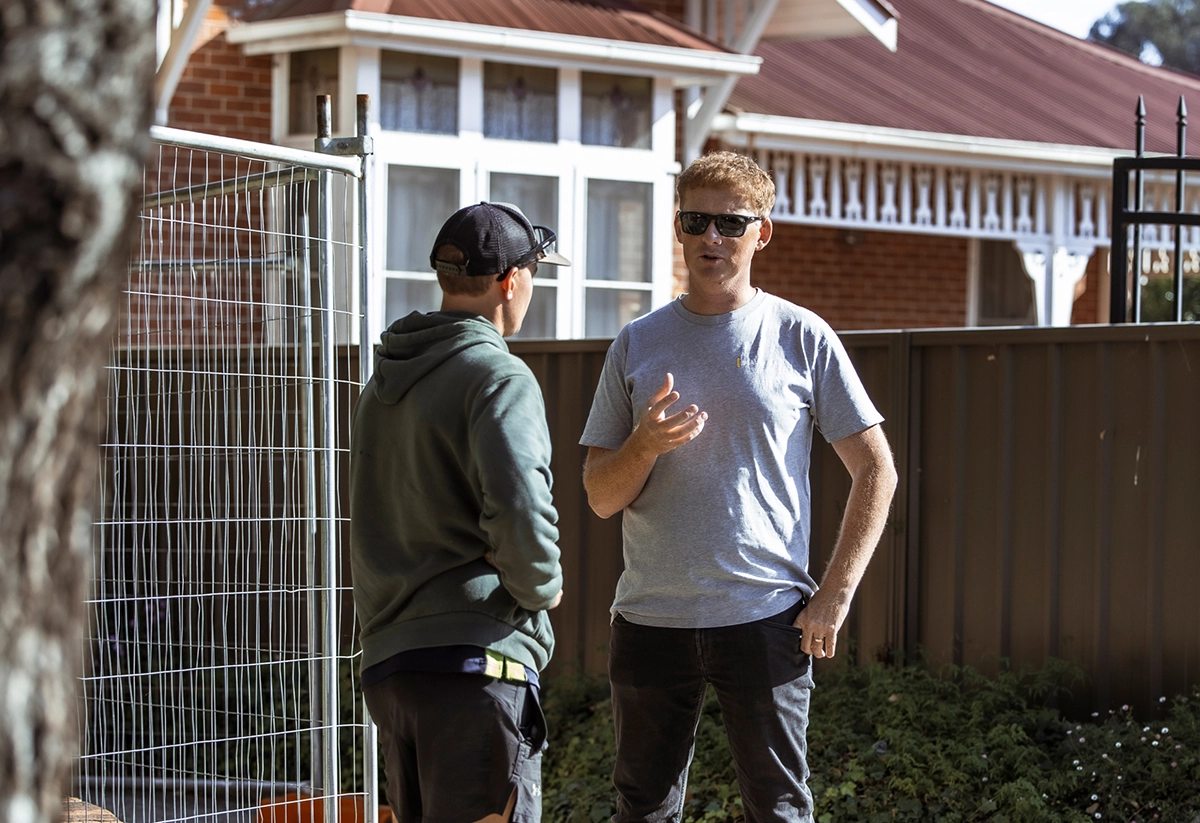 Two men discussing home improvement project in residential backyard with fencing installation visible.