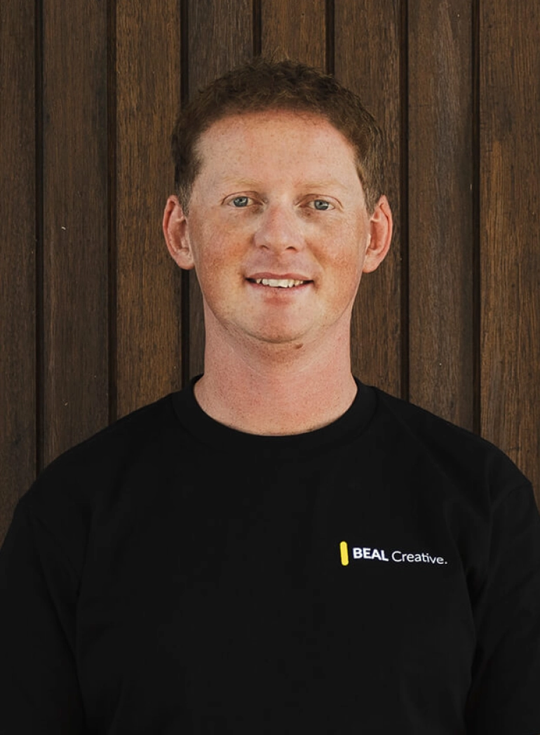 Man wearing black BEAL Creative shirt, smiling at camera against wooden backdrop. Web design and creative services.