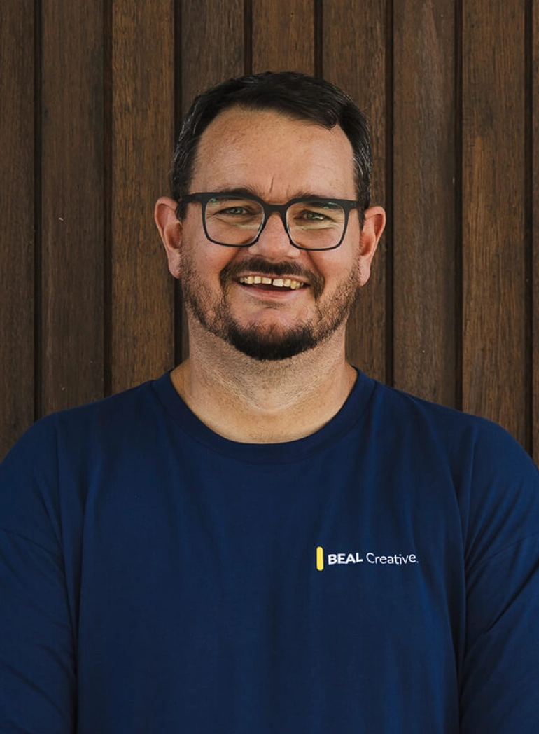 Man wearing BEAL Creative navy t-shirt, smiling at camera against wooden background. Web design and creative services.